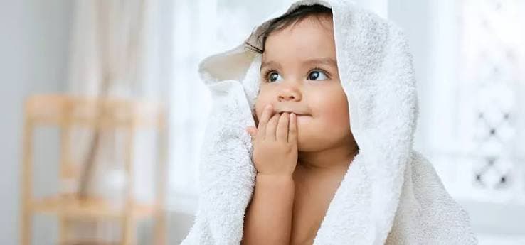 Bébé enveloppé dans une serviette blanche après le bain, la main près de la bouche, l’air curieux