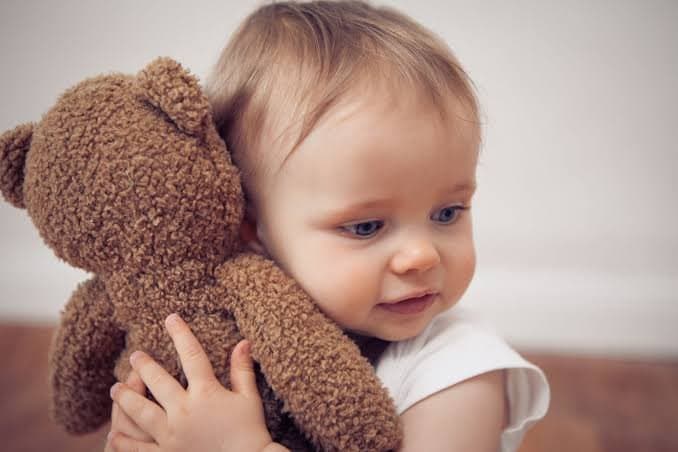 Un bébé blond aux yeux bleus tendre tendre contre sa joue un ours en peluche marron à bouclés poils.