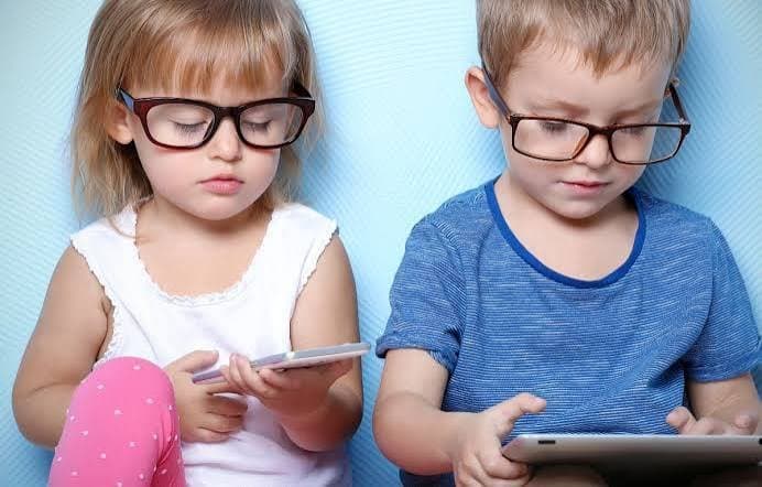 Deux jeunes enfants portant des lunettes, assis côte à côte et absorbés par un smartphone et une tablette